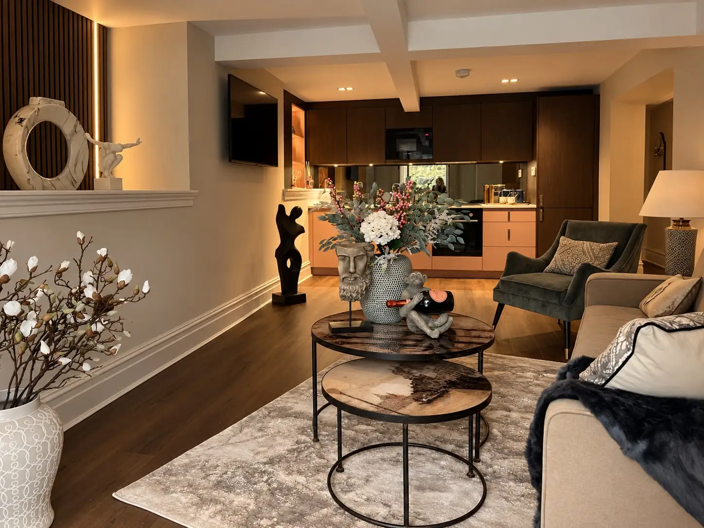 After: the furnished lounge with nesting coffee tables, a green armchair, sculpture and flowers in front of the kitchen