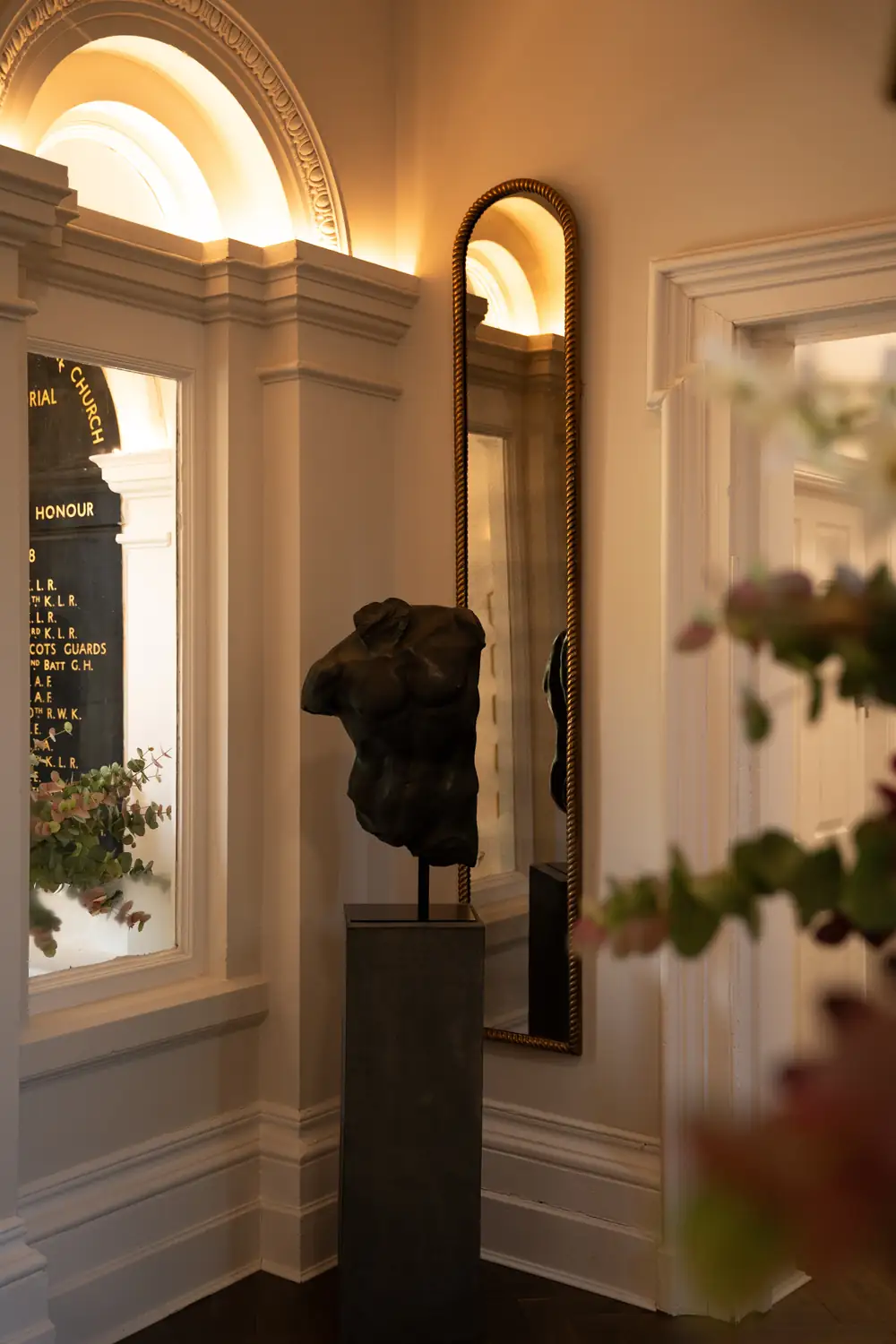 Dark torso sculpture on a plinth beside a tall arched mirror