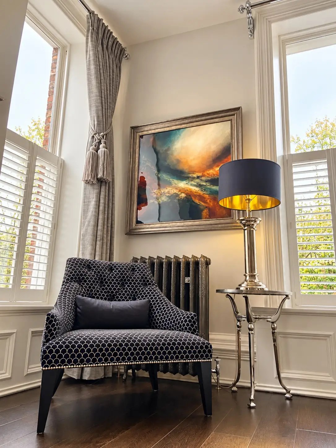Navy patterned armchair beside a brass lamp and a framed abstract painting, between two shuttered windows